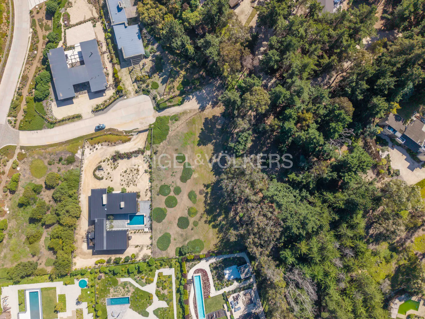 Aerial view of a property with modern homes, pools, and a large grassy lot bordered by trees. Engel & Völkers logo is visible.