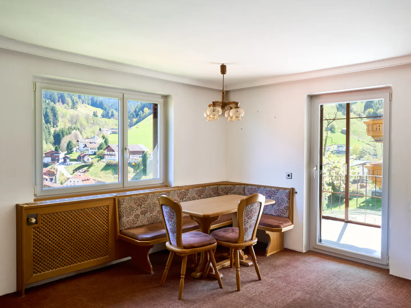 A dining area with a wooden table, chairs, and a corner bench. A window shows a view of green hills and houses.
