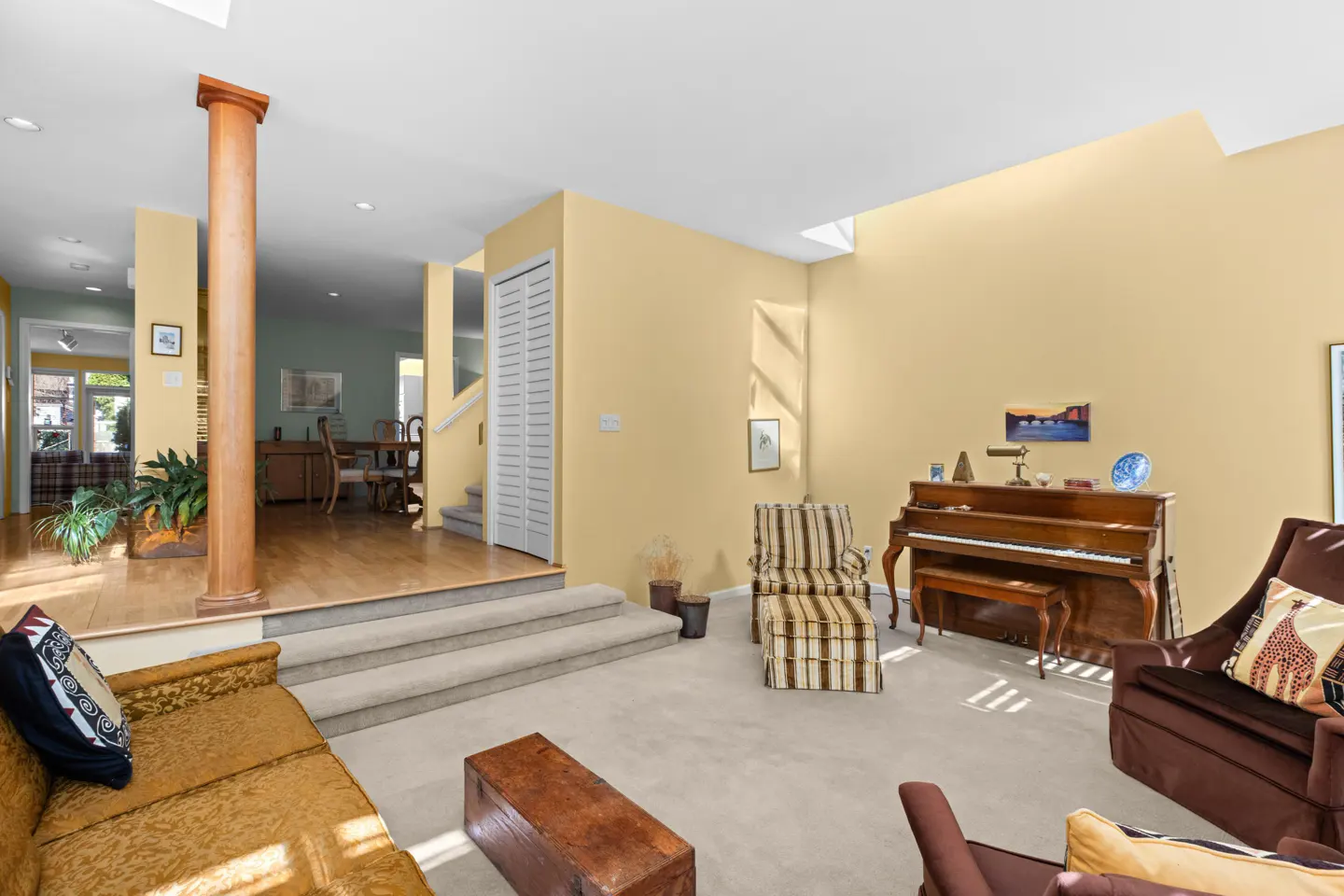 A living room with a piano, brown sofa, and striped chair. The walls are yellow, and there is a wooden pillar.