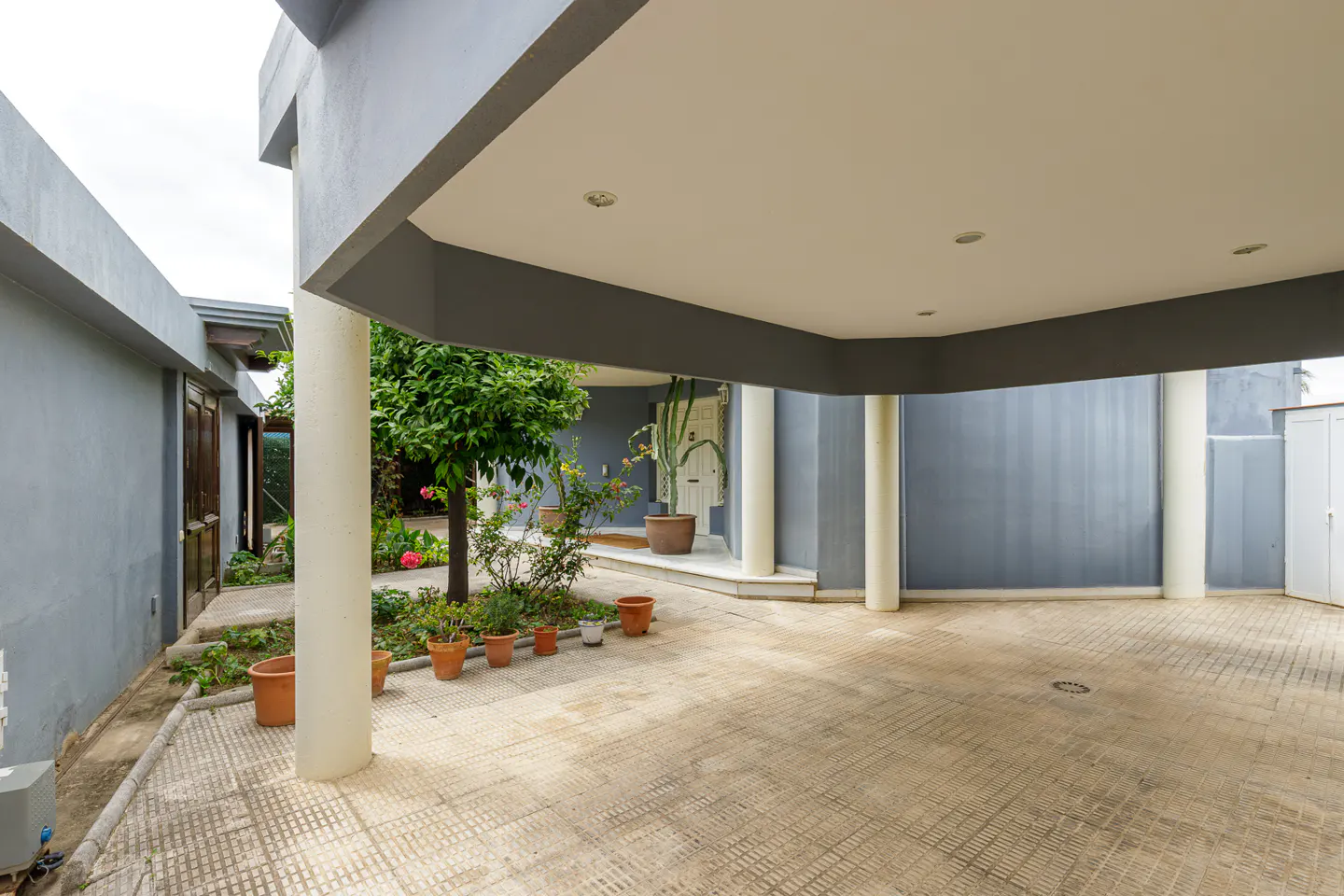 Covered patio with gray walls, white columns, and a tiled floor. Potted plants and a tree add greenery. A white door is visible in the background.
