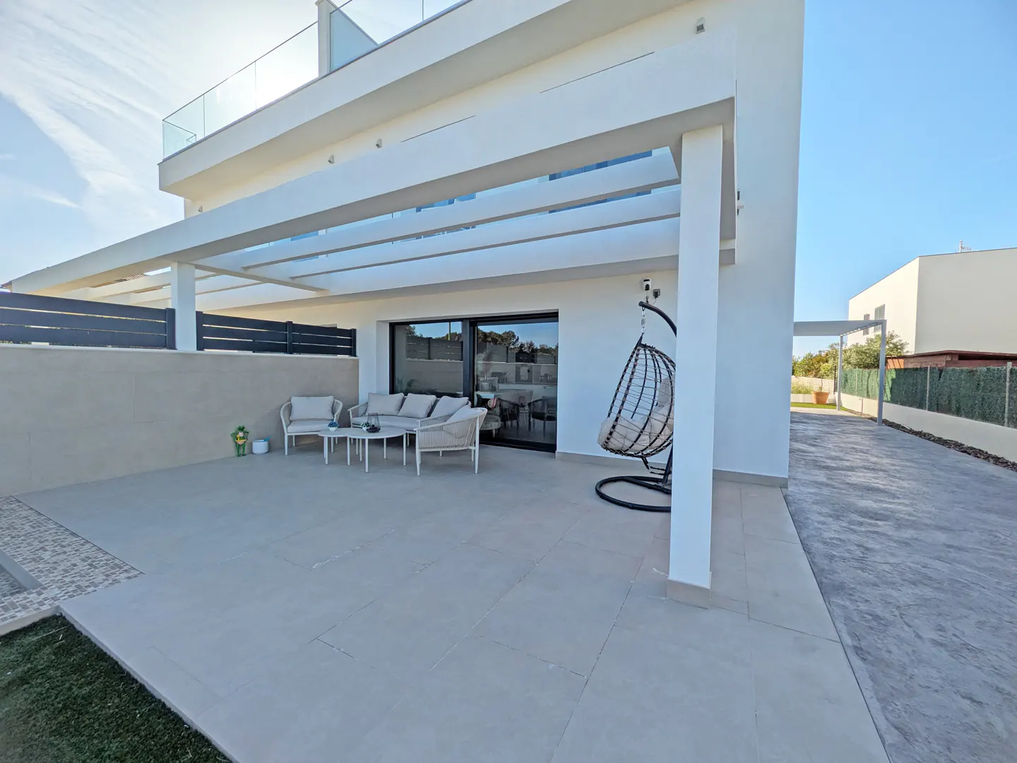 Modern white house exterior with patio furniture, hanging chair, and a pergola.