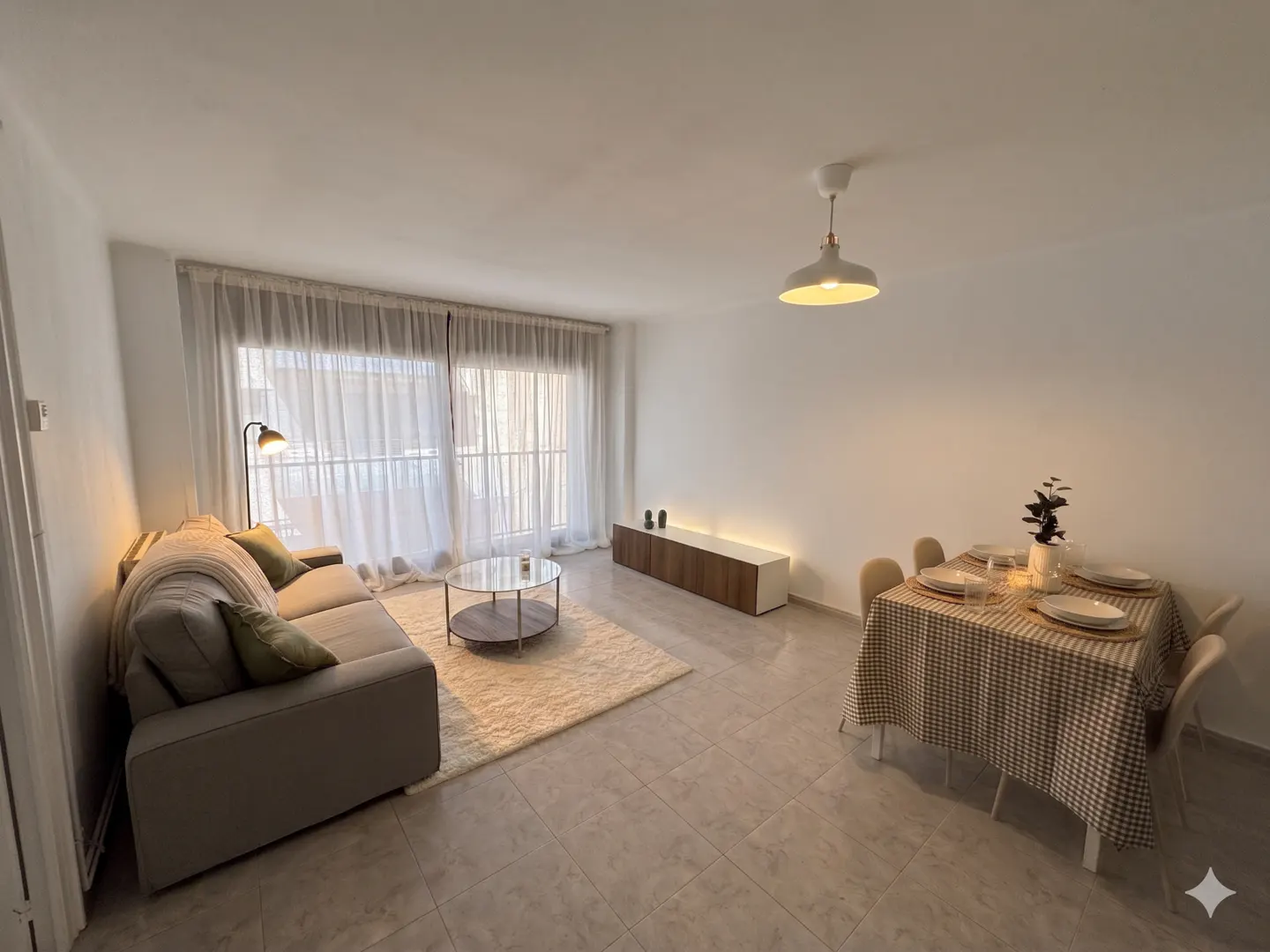 Bright, open-plan living and dining area with a gray sofa, round glass table on a cream rug, and a dining table set for four.