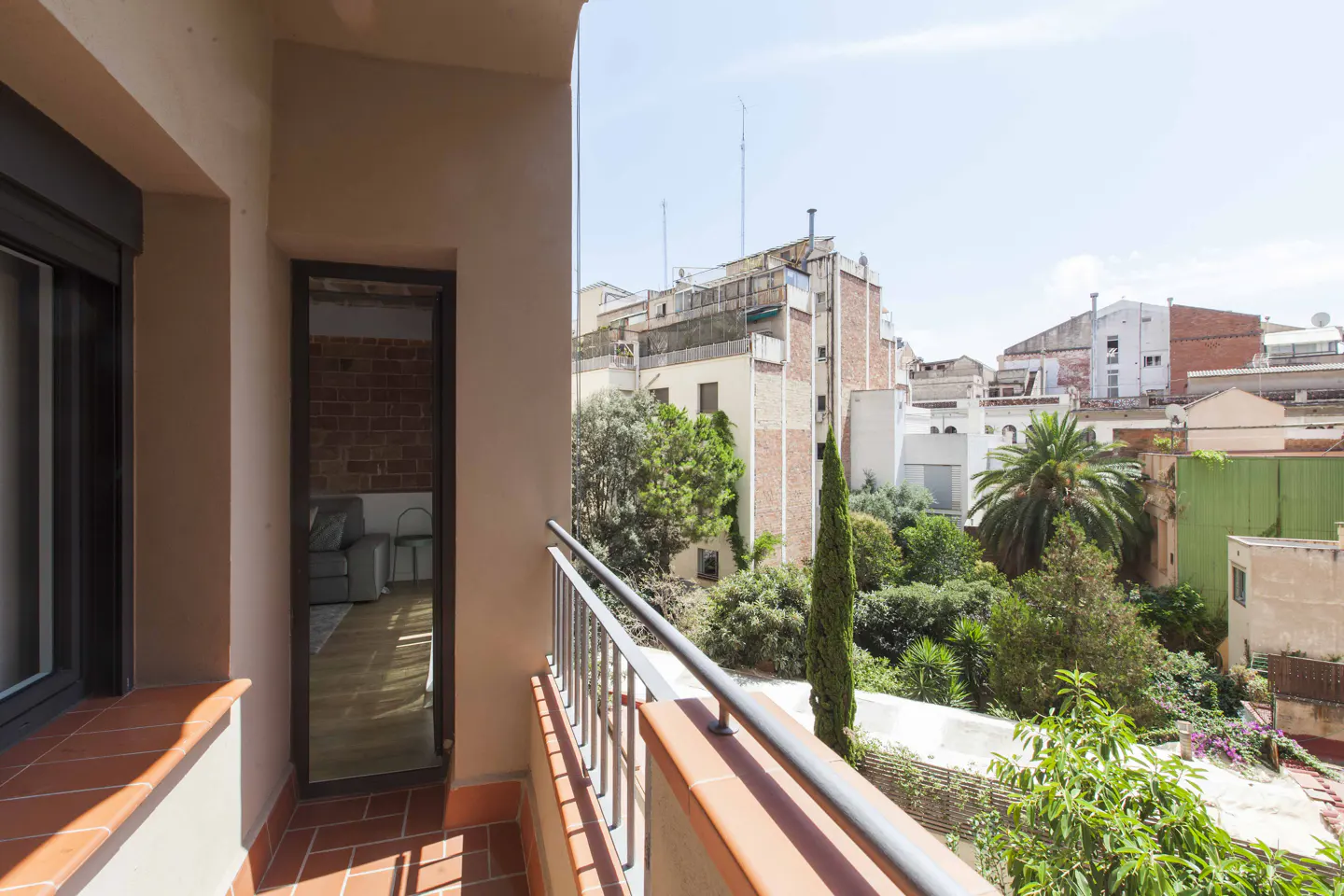 A balcony with terracotta tiles and a metal railing overlooks a lush green garden and urban buildings under a clear sky.