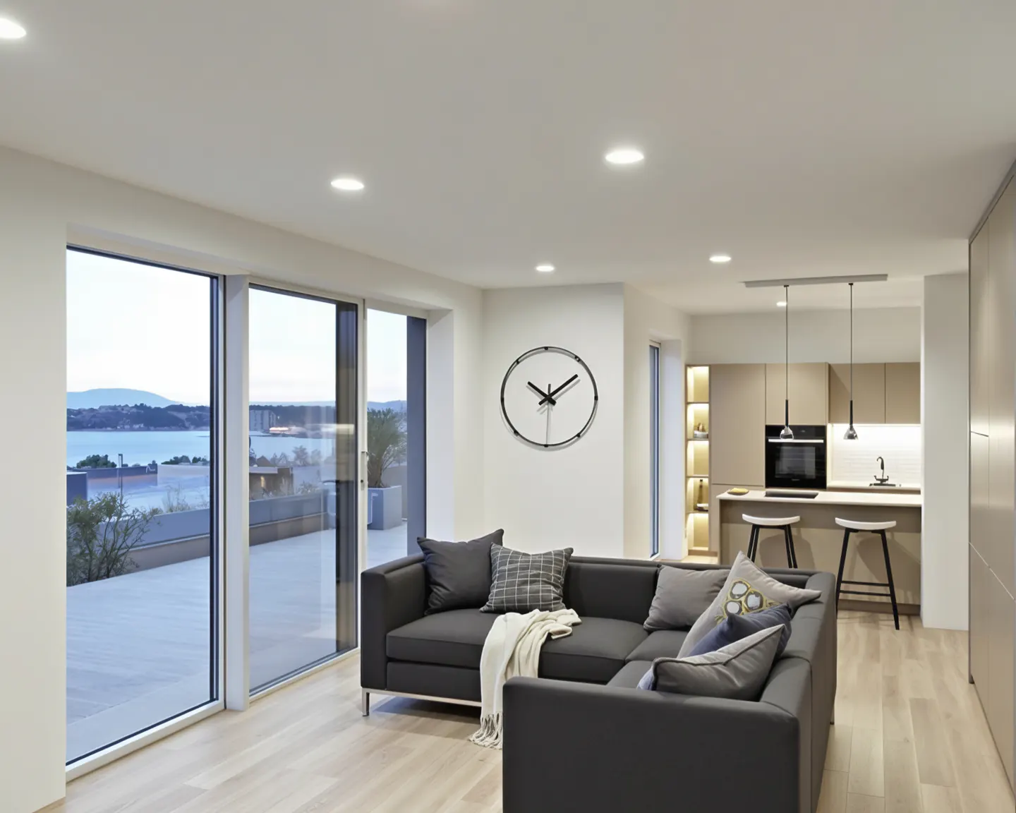 Modern open-plan living room with a dark grey sectional sofa, large wall clock, and kitchen island overlooking the ocean.