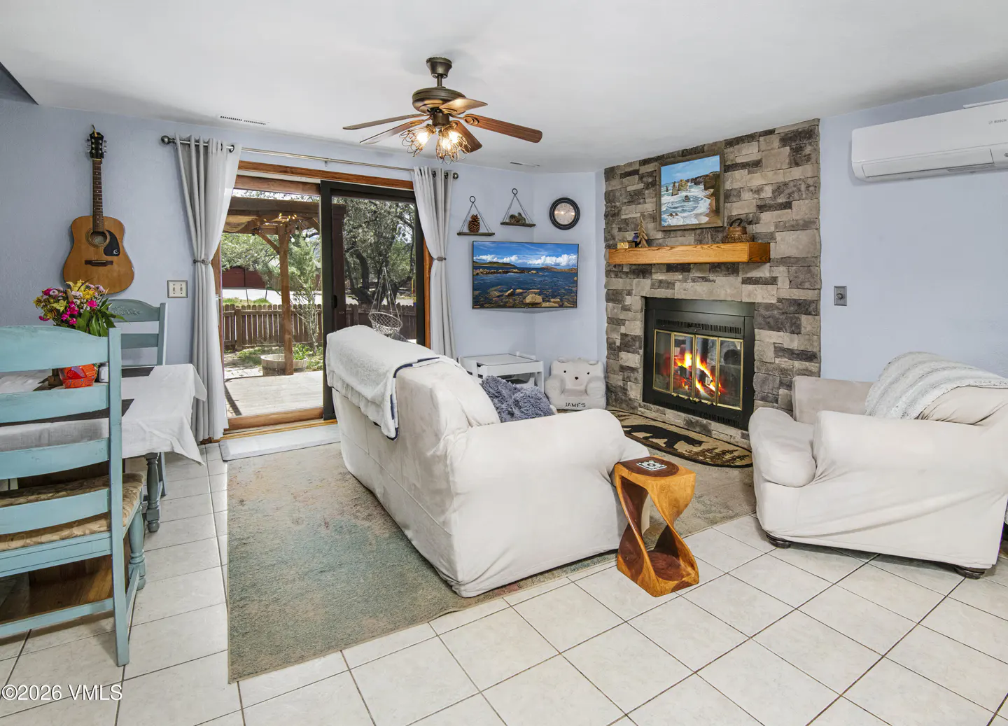 Living room with white tile floor, stone fireplace, and white slipcovered furniture. A guitar hangs on the blue wall near a dining table. Sliding glass doors lead to a patio.