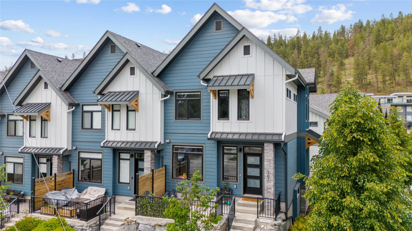 Exterior view of blue and white townhouses with gray roofs, black trim, and small front patios. A wooded hillside is in the background.