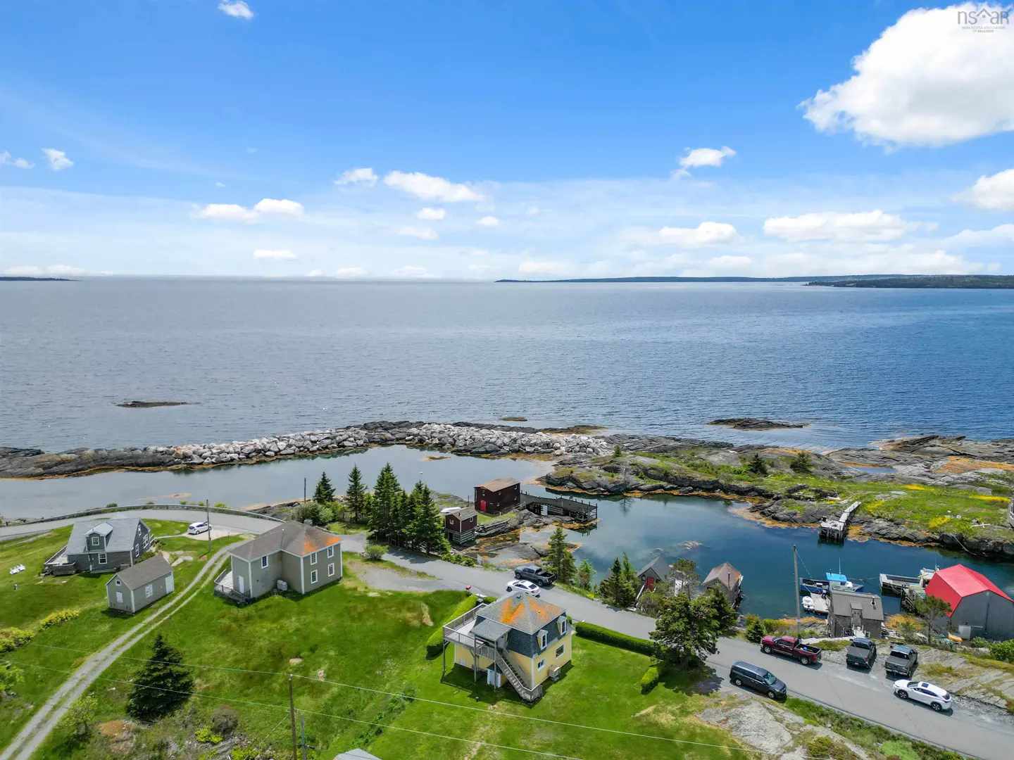 Aerial view of coastal homes near a rocky shoreline and calm blue ocean under a bright, partly cloudy sky.
