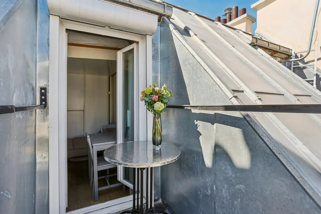Balcony with a metal table holding a vase of flowers. An open door leads to a small room with a table and chairs.