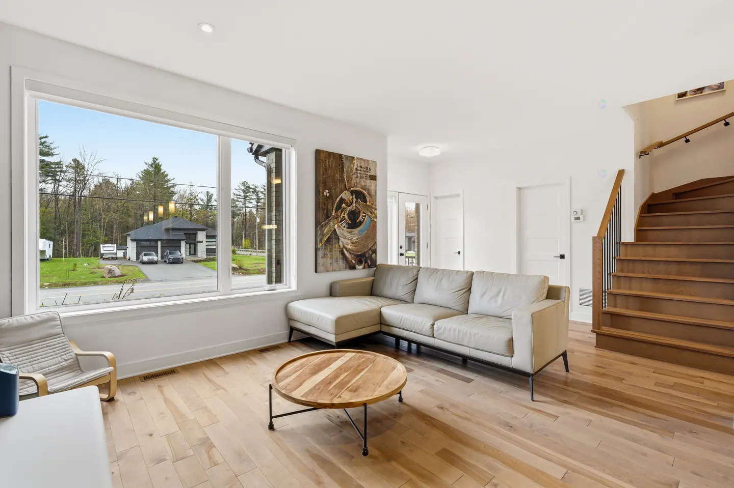 Bright living room with hardwood floors, a beige sectional sofa, and a round wooden coffee table. A large window overlooks a modern house.