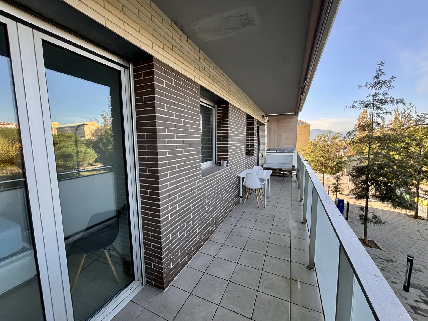 A balcony with a brick wall, a white table and chairs, and a view of trees and a street.