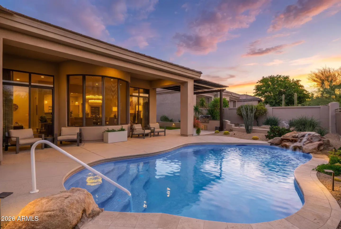 Backyard pool at dusk with a waterfall feature, patio furniture, and desert landscaping.