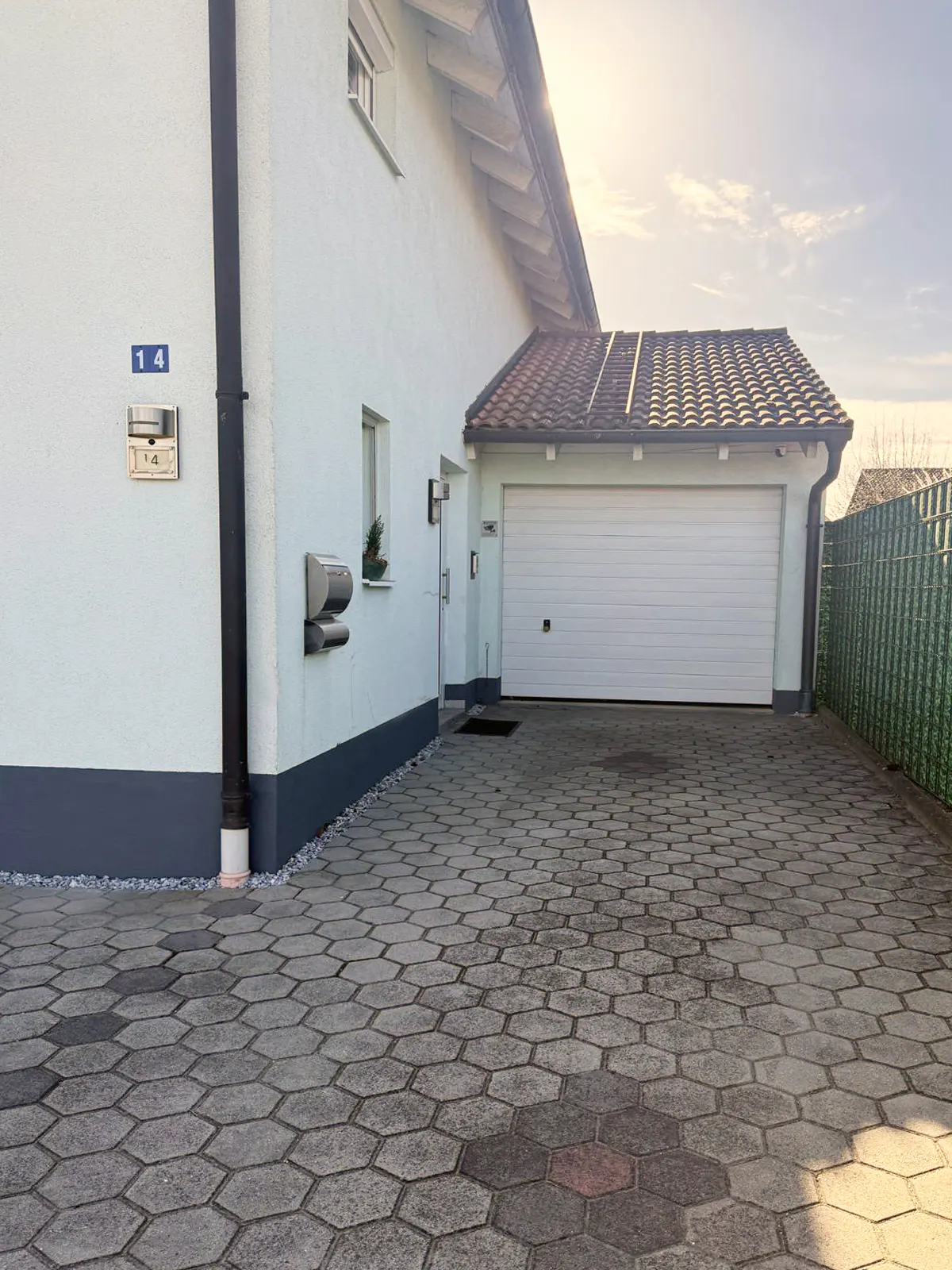 Exterior view of a light blue house with a white garage door and a gray hexagonal-tiled driveway.