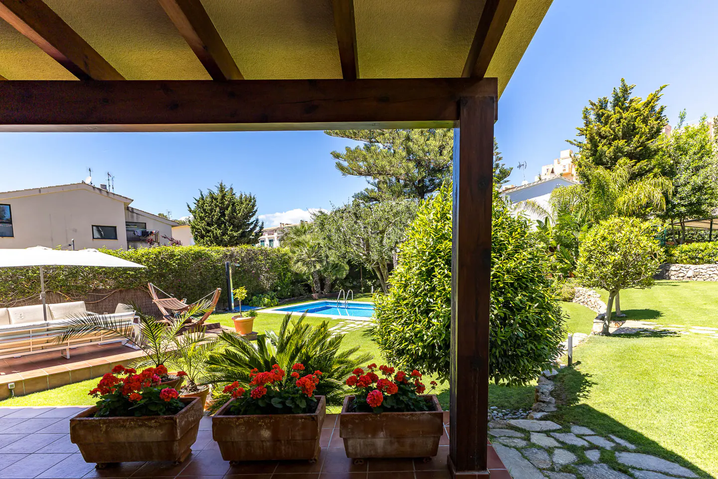 View from a covered patio with red flowers in pots, overlooking a green lawn, pool, and trees on a sunny day.