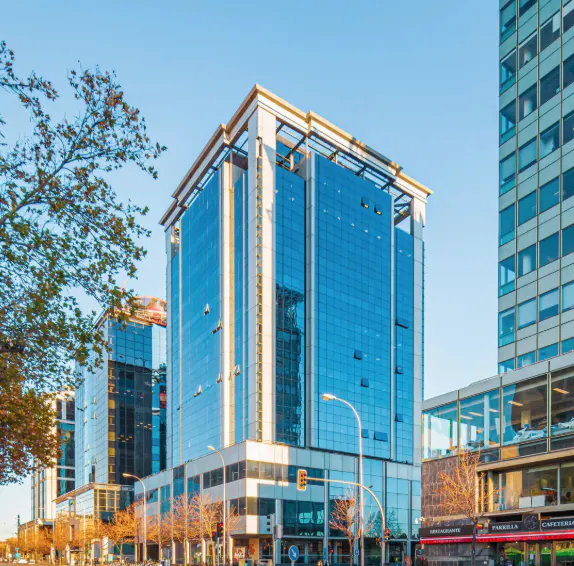 Modern blue glass office building on a city street with trees and shops.