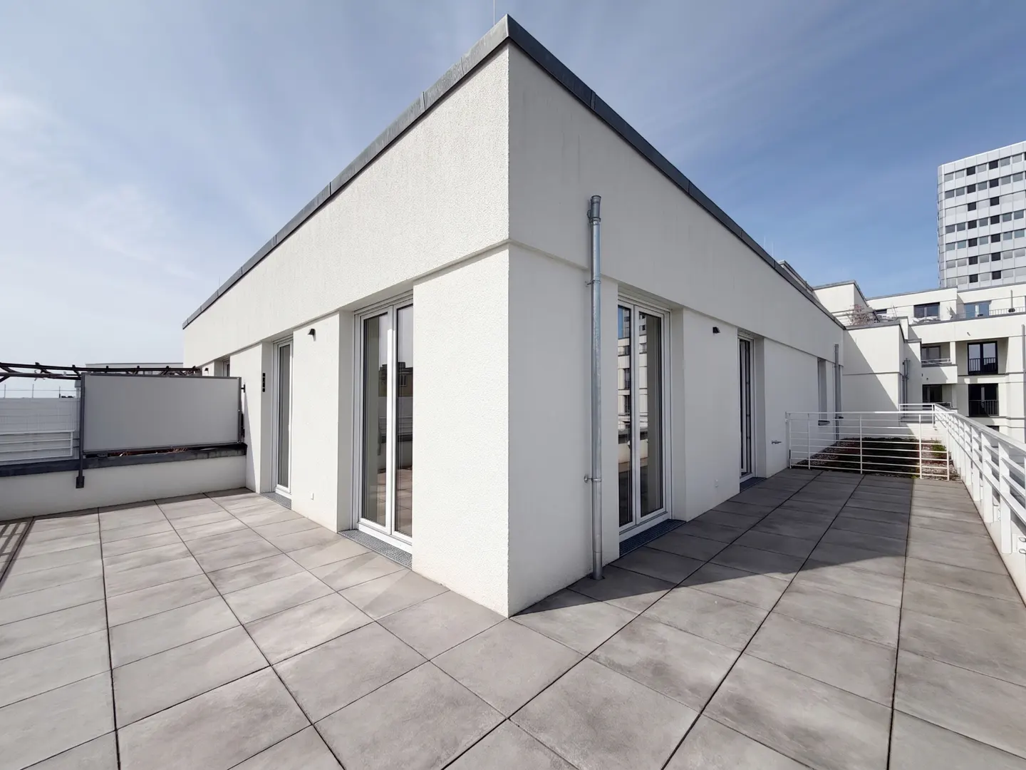 Wide, sunlit rooftop terrace with gray tile flooring and white building exterior. Sliding glass doors lead inside. Railings line the perimeter.