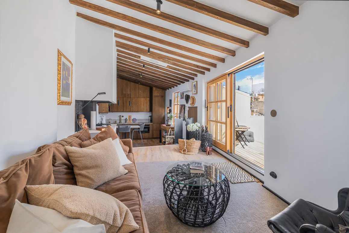 A bright living room with a brown sofa, a black coffee table, and a kitchen in the background. The room has a wooden beamed ceiling and a sliding door to a balcony.