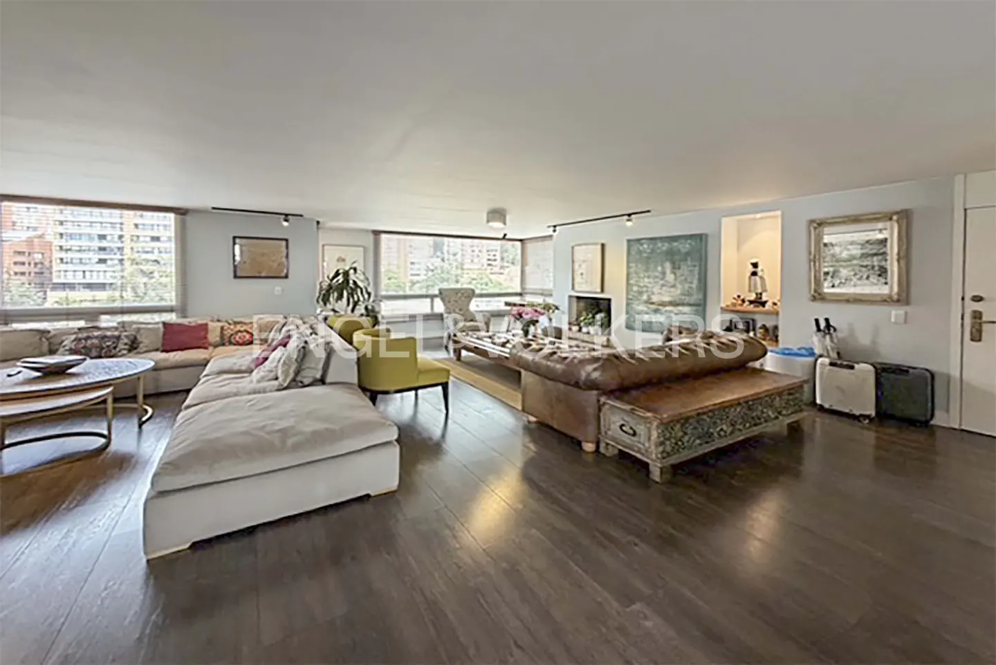 A spacious living room with a large sectional sofa, leather couch, and dark wood floors. Windows overlook a city.