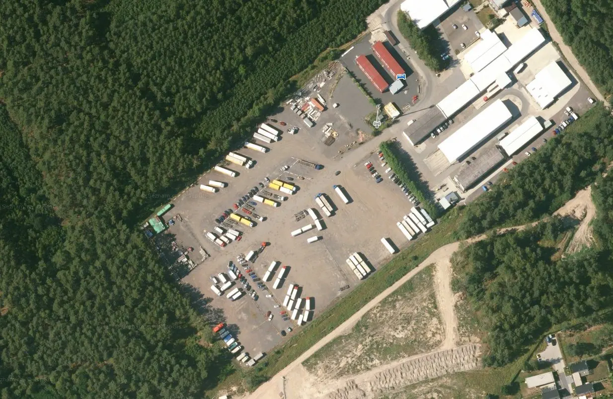 Aerial view of a commercial property with white buildings, a parking lot filled with trucks, and a dense green forest.