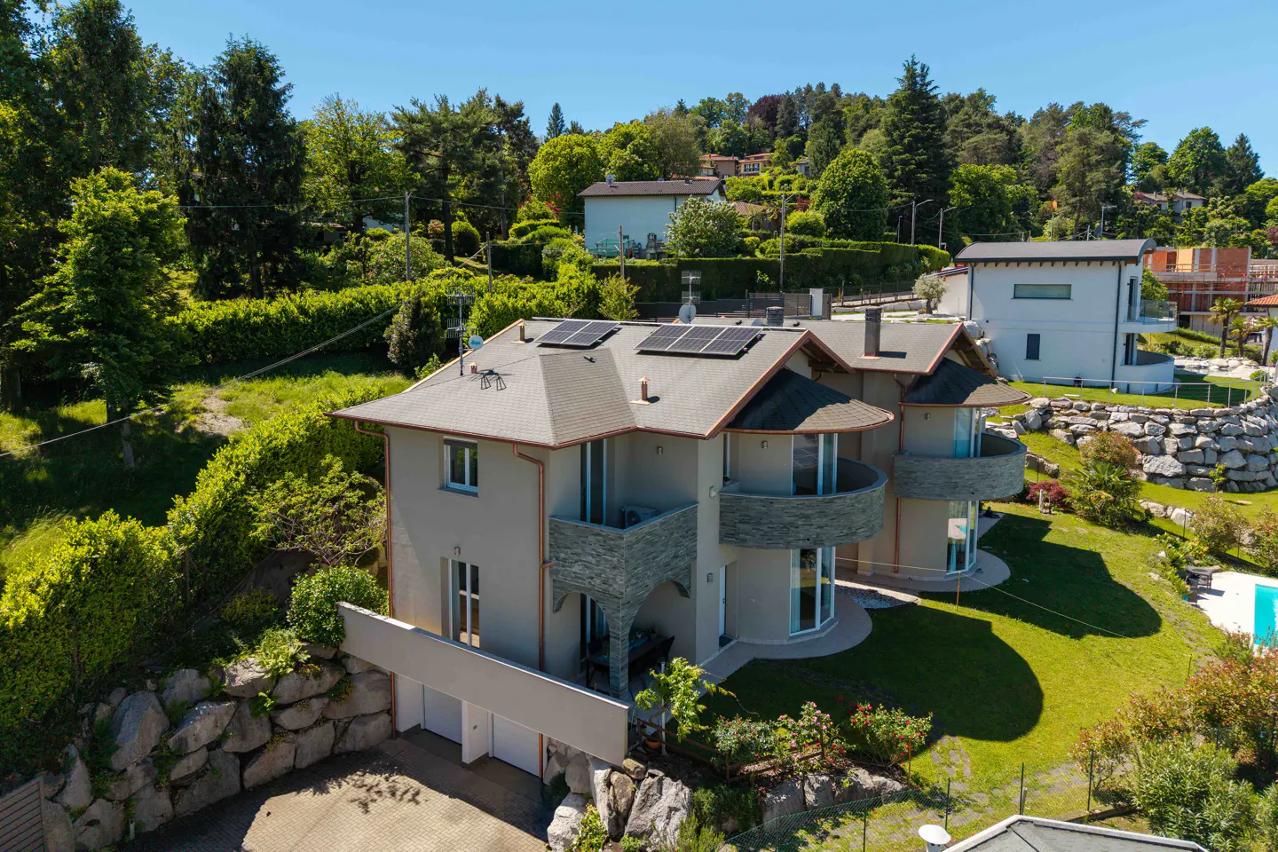 Aerial view of a modern, multi-story house with a gray exterior, stone accents, and solar panels on the roof, surrounded by lush greenery.