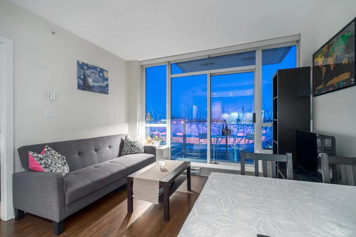 Living room with gray sofa, coffee table, and dining table. Large windows show a night view of a harbor. "Starry Night" art on the wall.