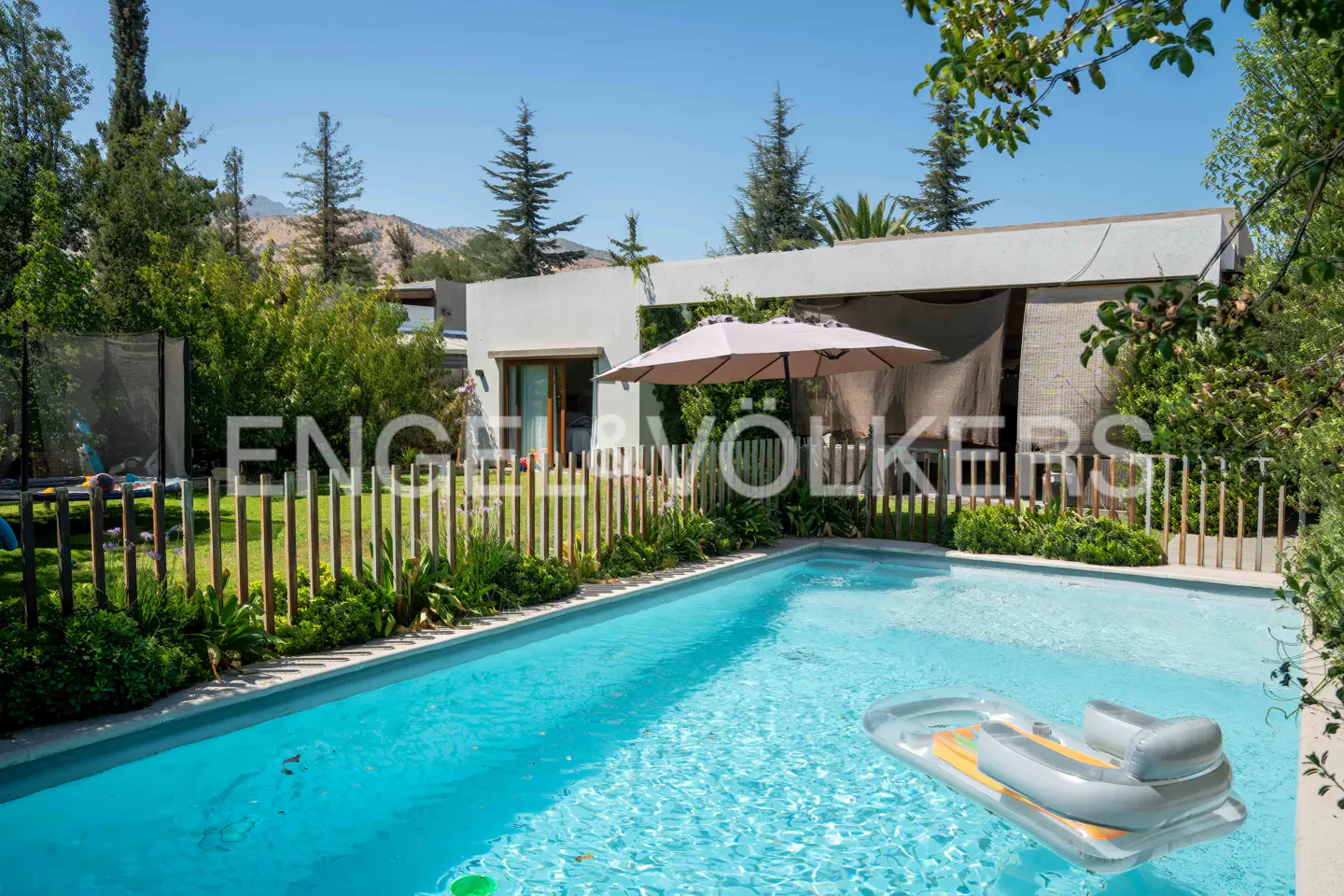 Outdoor pool with a gray inflatable raft, surrounded by a wooden fence, green plants, and a house with a beige umbrella.