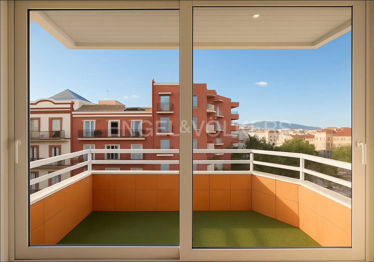 View from a balcony with green turf, orange walls, and white railings, overlooking red buildings and a city skyline under a blue sky.