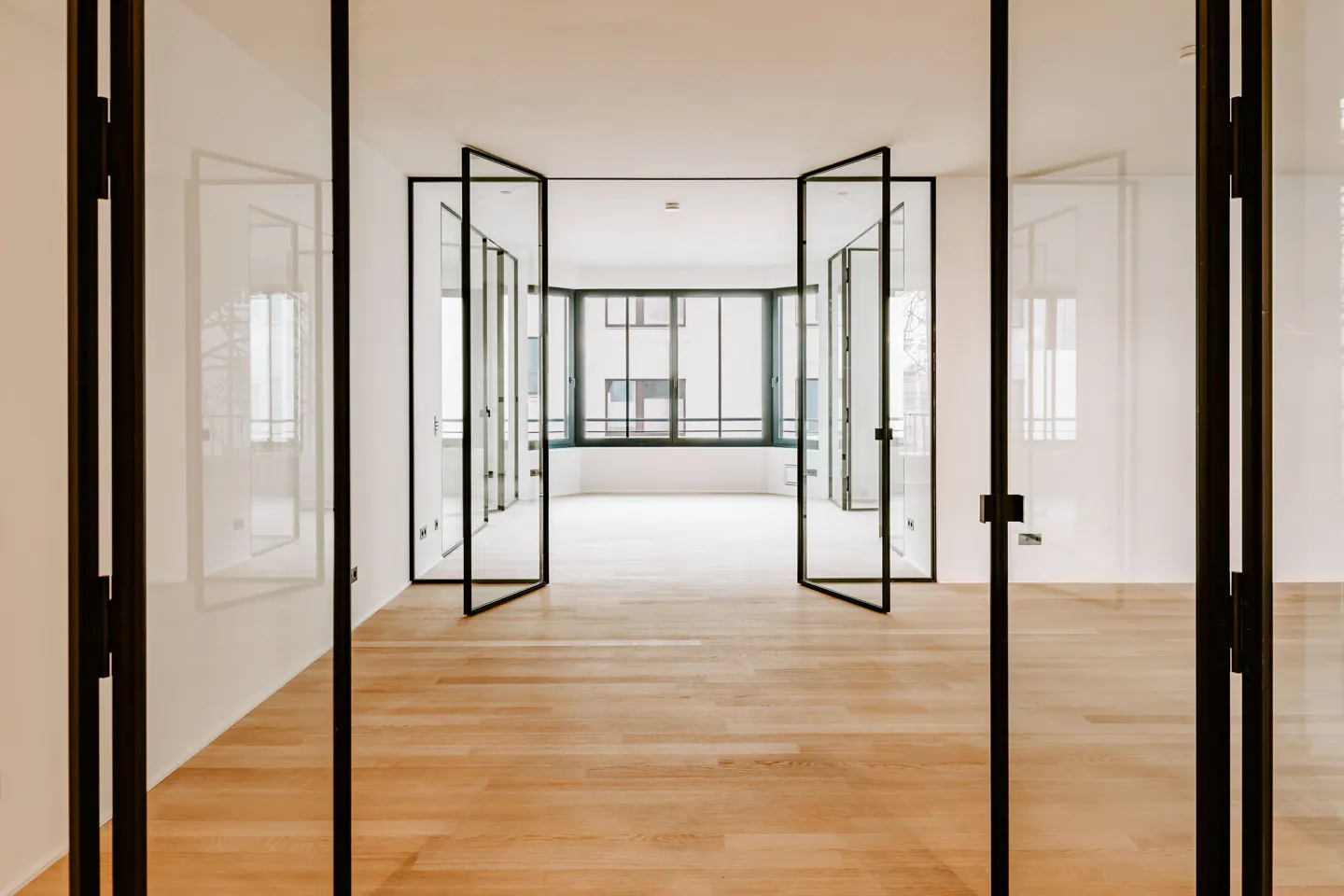 Interior view of a bright, empty room with wood floors and white walls. Black-framed glass doors are open, leading to a window with a city view.