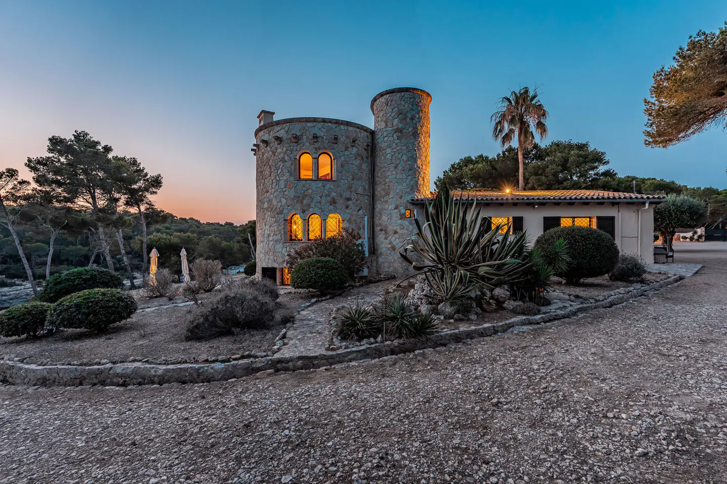 Stone castle-like home with a tower, lit windows, and lush landscaping at dusk.