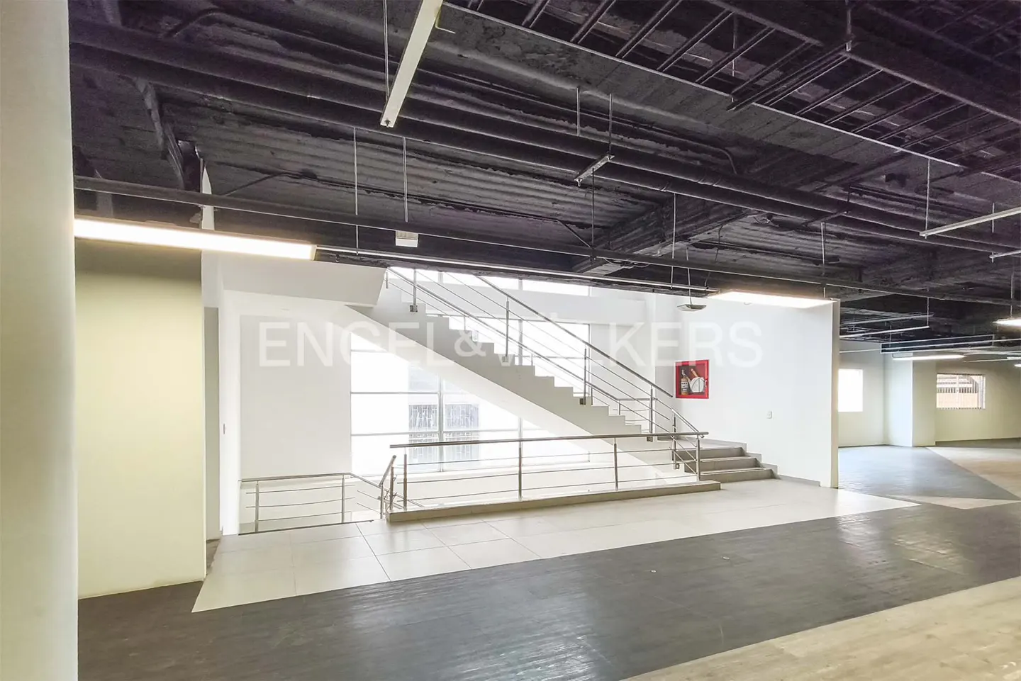 Interior view of a commercial space with stairs, metal railings, and a red fire extinguisher on a white wall.