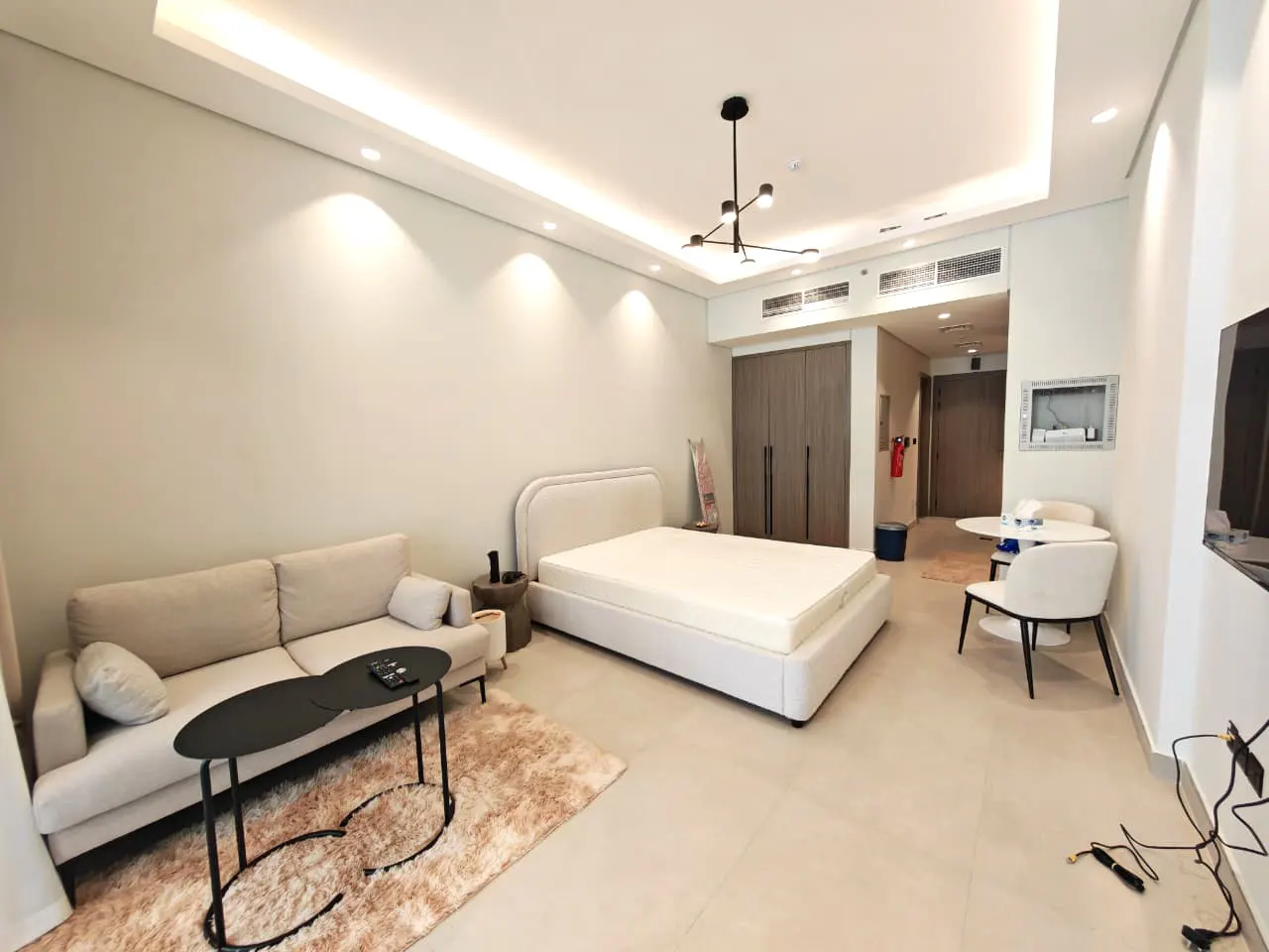 A bright studio apartment with a beige sofa, black coffee table, and a white bed. A small dining set is visible in the background.