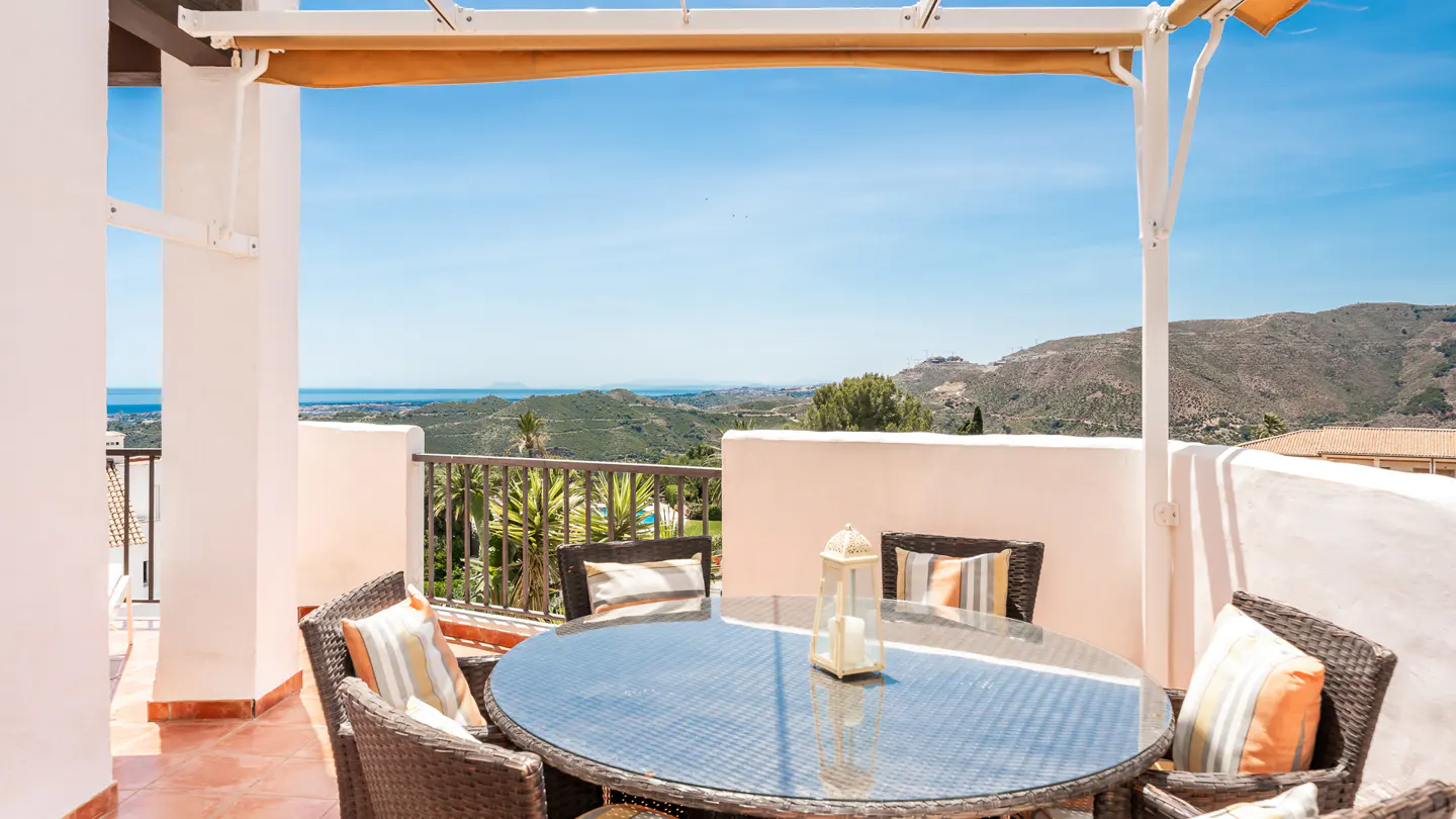 Outdoor patio with wicker furniture, a glass table, and a lantern. A scenic view of mountains and the ocean is in the background.