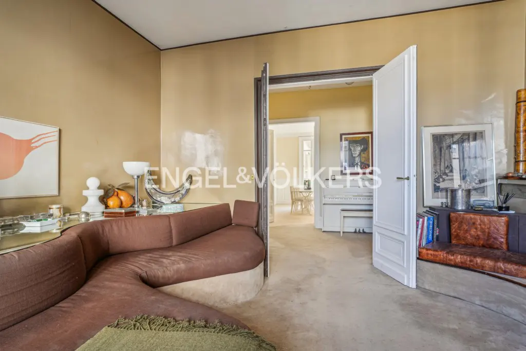 Living room with brown curved sofa, tan walls, and open doorway to a room with a white piano. Artwork and decor are visible.