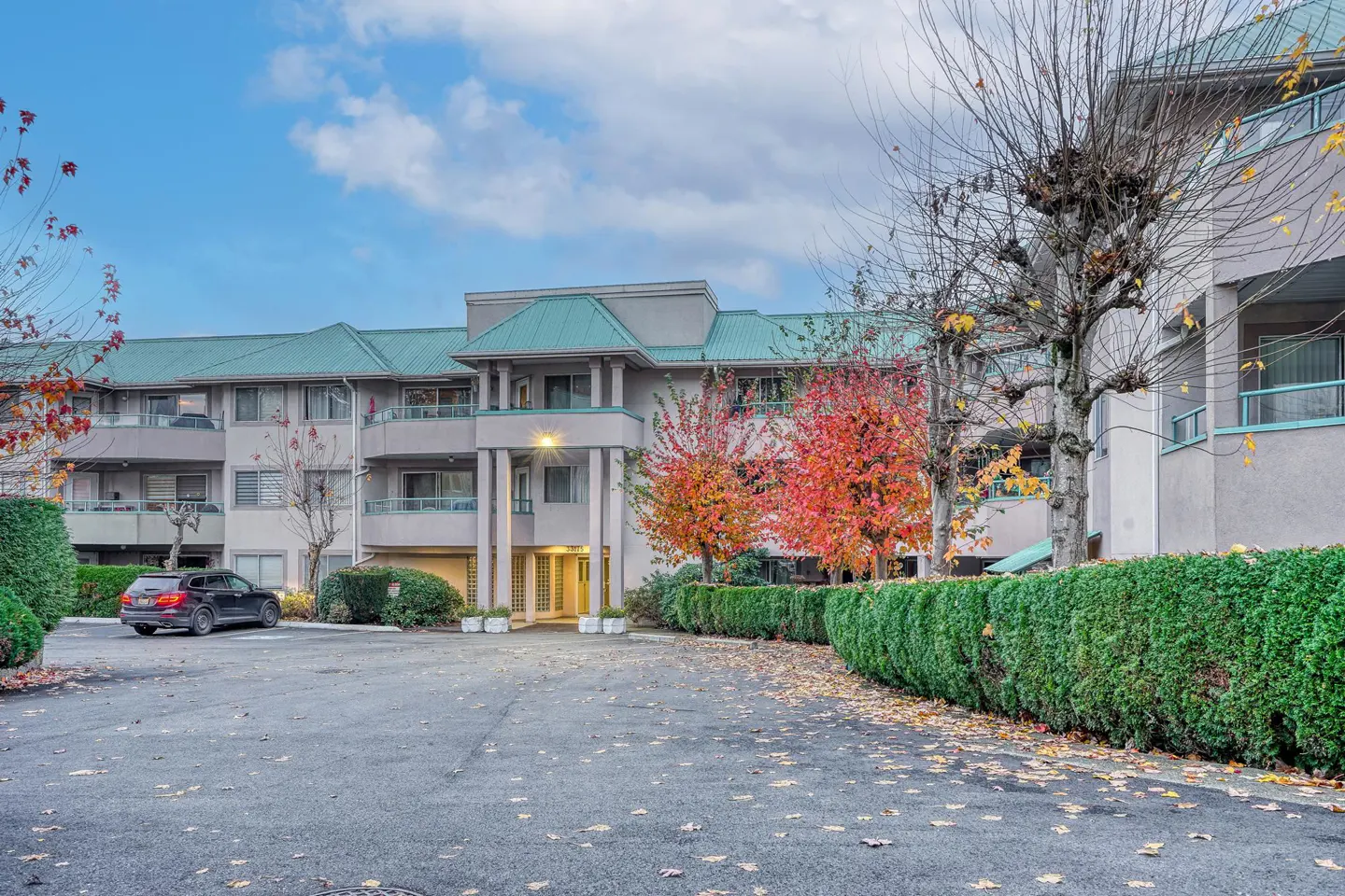 Exterior view of a three-story apartment building with a green roof and balconies, surrounded by trees and bushes.