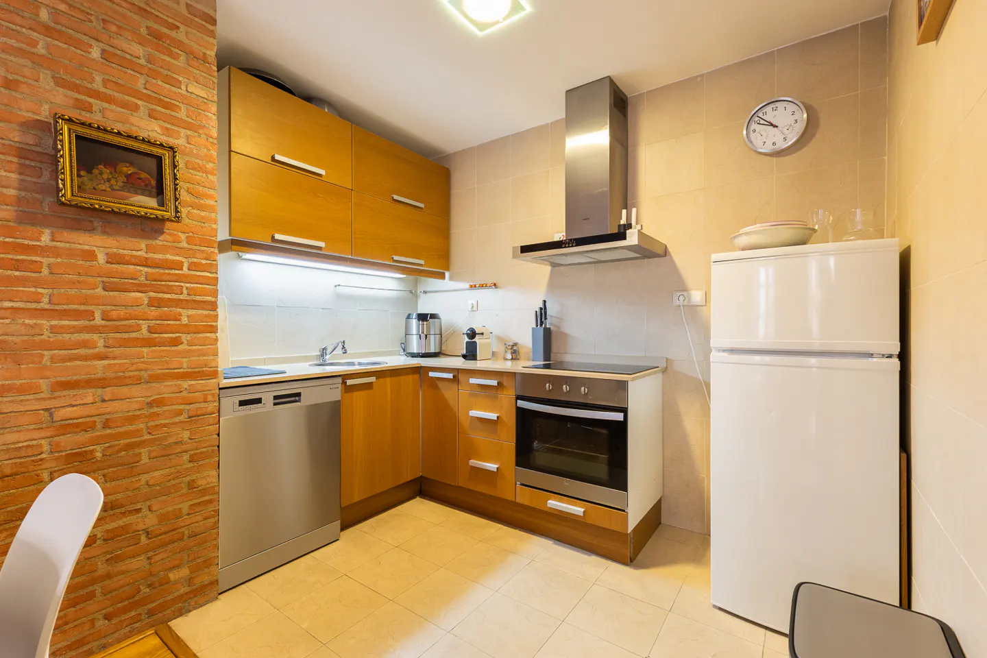 A kitchen with light wood cabinets, stainless steel appliances, and a white refrigerator. A brick wall with a framed painting is on the left.