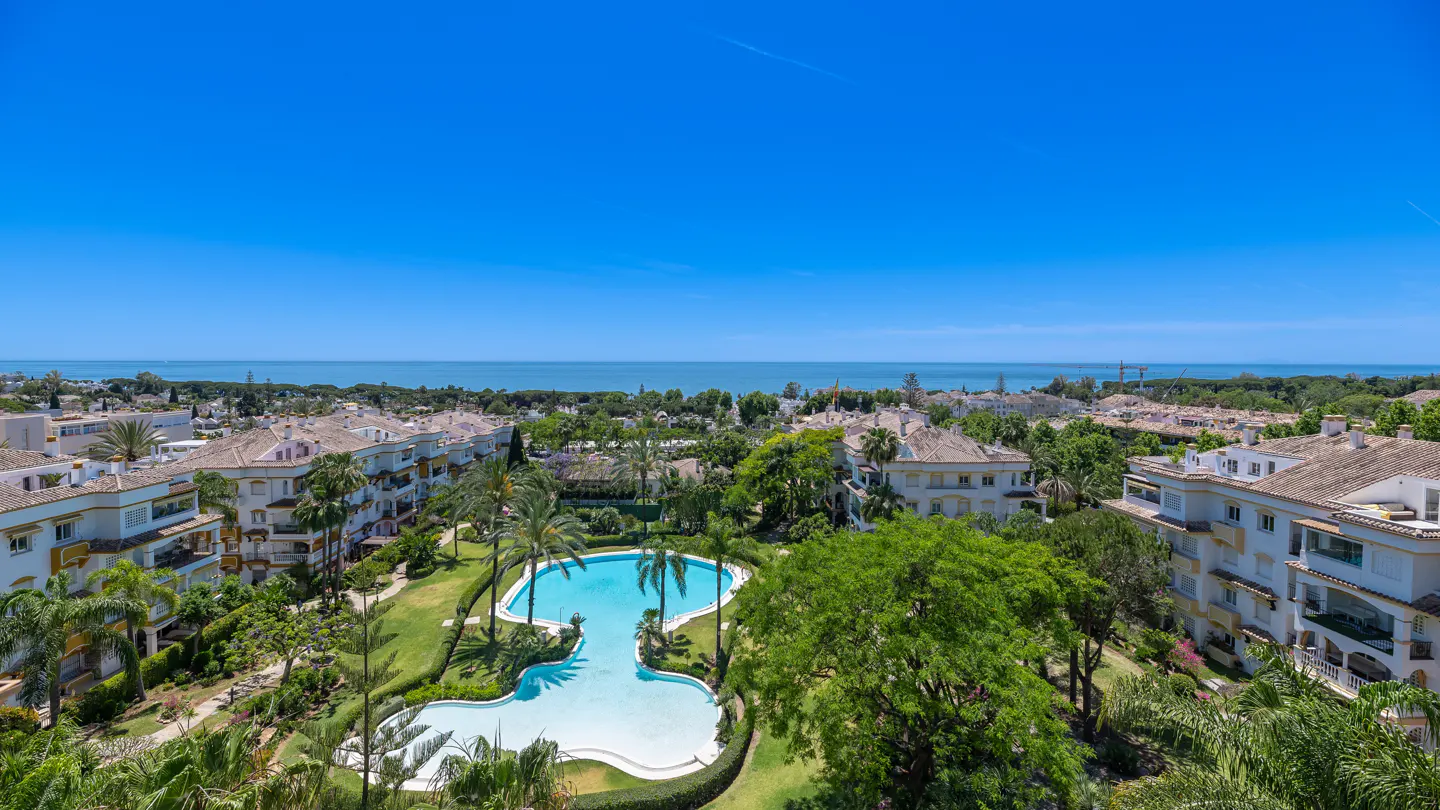 Aerial view of a resort with a blue pool, palm trees, and white buildings with terracotta roofs under a clear blue sky.