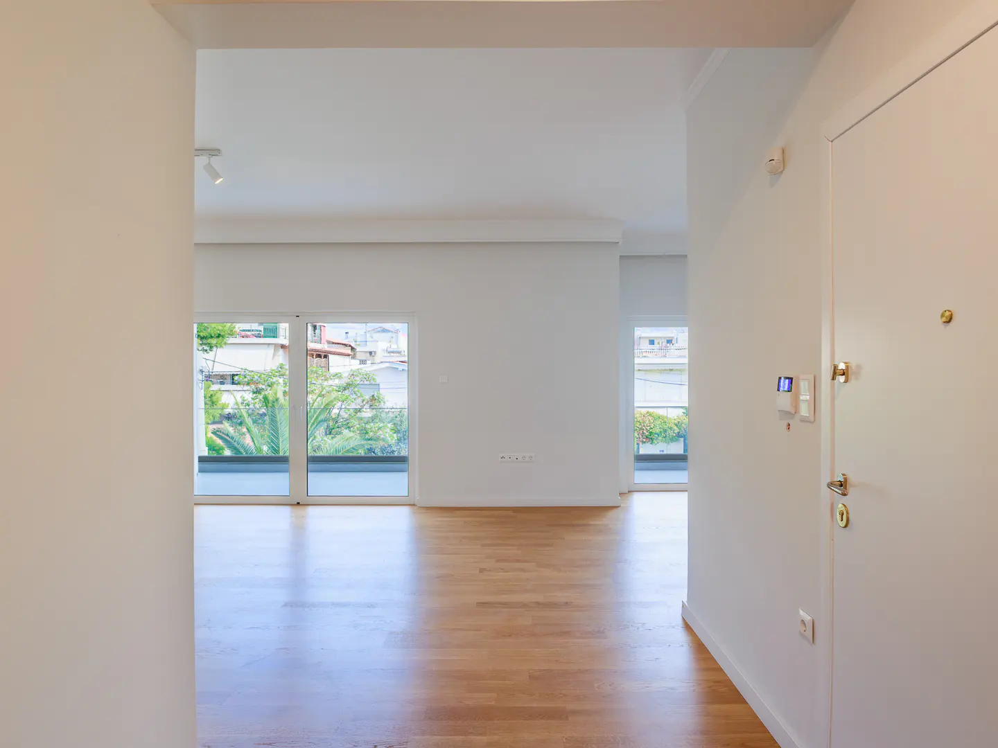 A bright, modern apartment entrance with a white door, light wood floors, and large windows showing green trees.