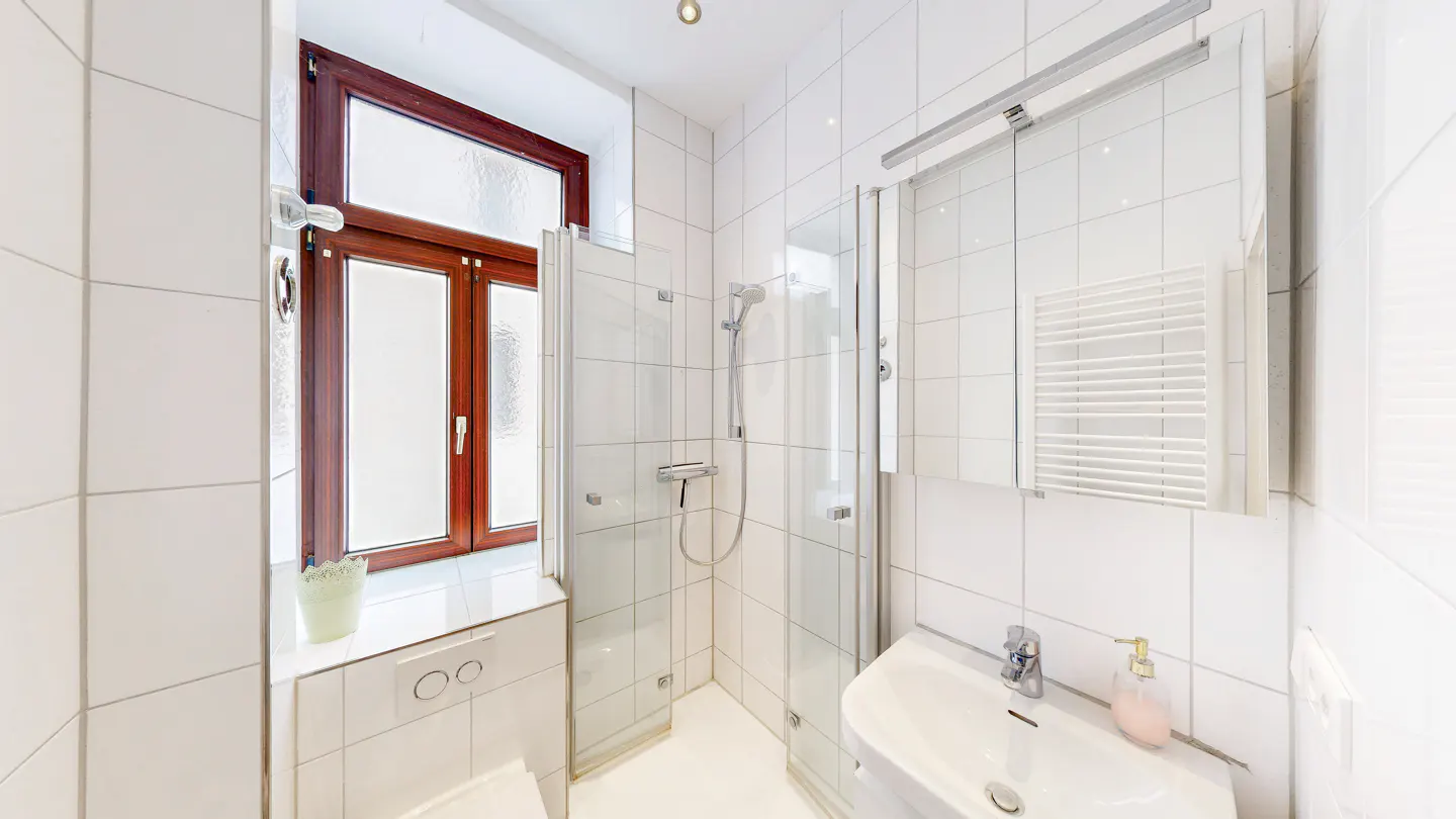 Bright bathroom with white tiles, a glass shower, and a white sink. A brown-framed window is on the left.