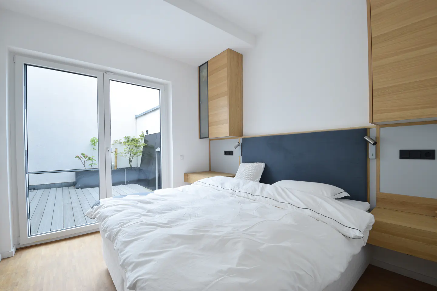 Bright bedroom with white bedding, a blue headboard, and a sliding glass door to a patio with plants.