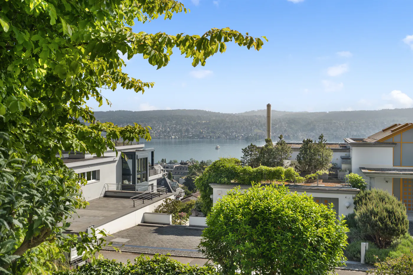 View of modern white homes with green foliage overlooking a lake and distant hills under a blue sky.