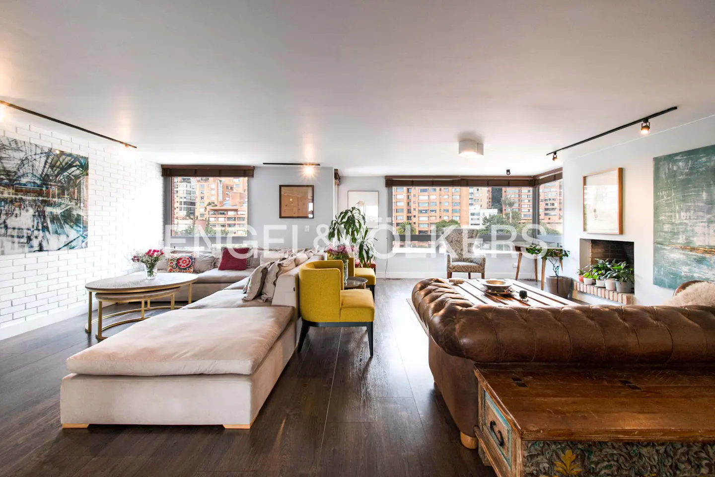 Bright living room with a large beige sectional, yellow chairs, and a brown leather sofa on dark wood floors. Windows overlook city buildings.