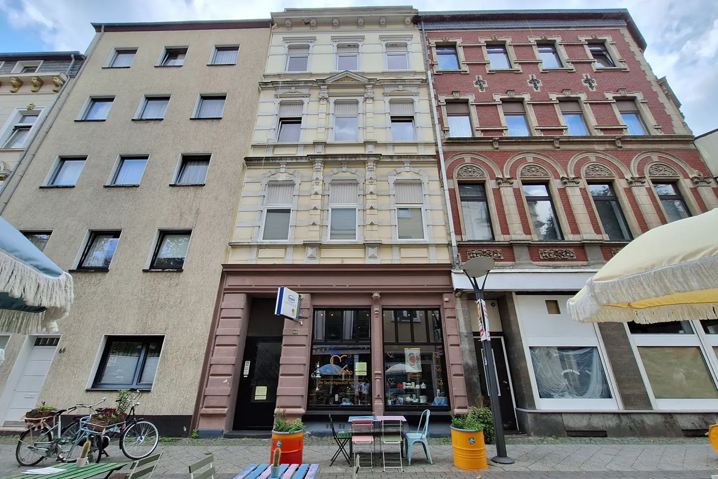 Street view of three buildings with different facades. The left building is beige, the middle is yellow, and the right is red brick. Bicycles and tables are on the sidewalk.