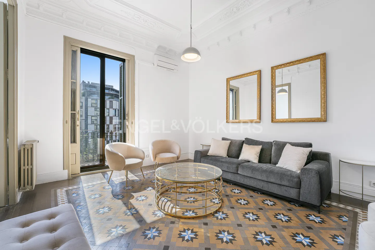 Bright living room with a gray sofa, two pink chairs, and a gold coffee table on a patterned tile floor. Two gold-framed mirrors hang on the white wall.
