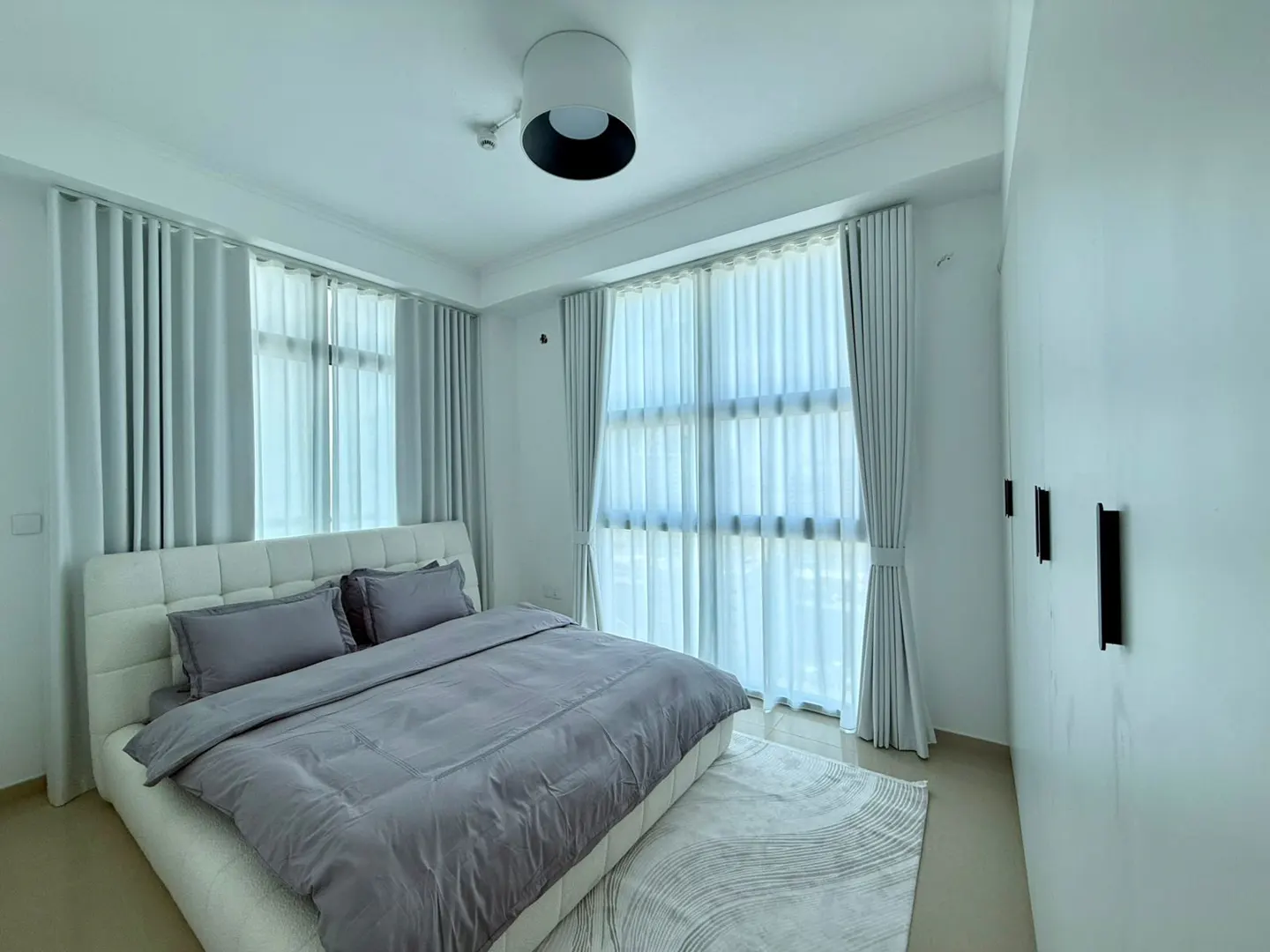 Bright bedroom with a white bed, gray bedding, and sheer curtains. A white rug sits on the floor, and a modern light fixture hangs from the ceiling.