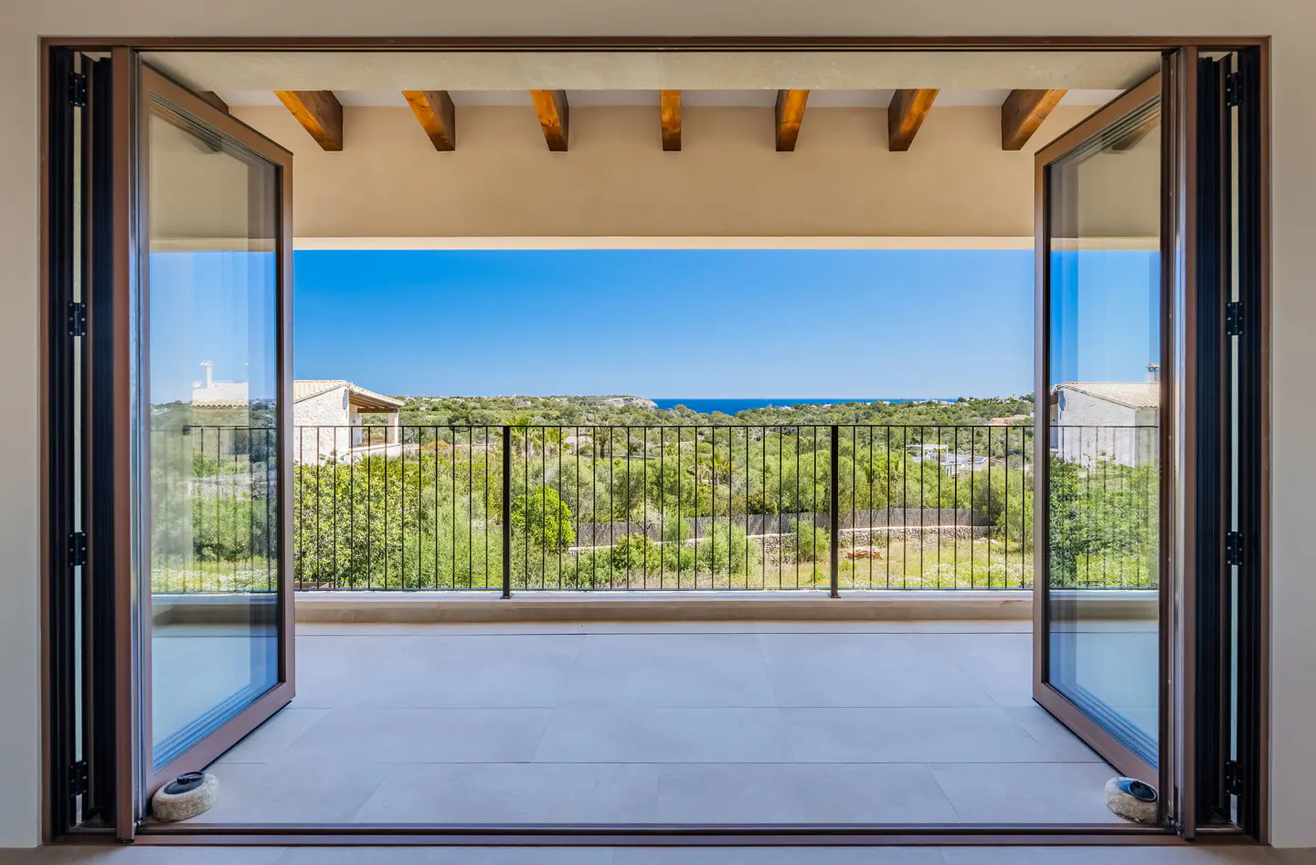 Open bi-fold doors lead to a balcony with a black railing, overlooking lush greenery and the blue sea under a clear sky.