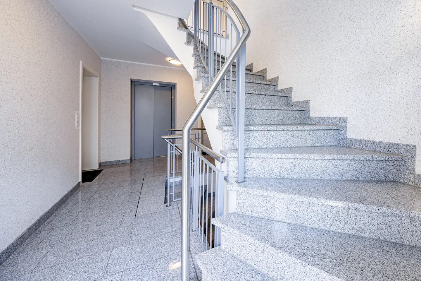Interior view of a building's staircase with gray granite steps and stainless steel railings, plus an elevator.
