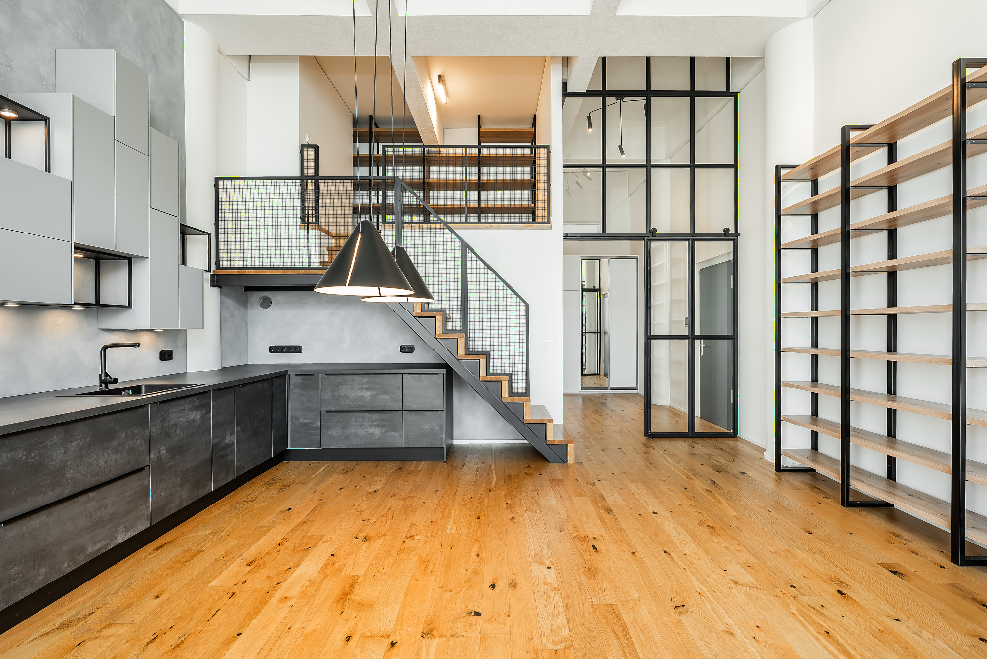 Modern loft kitchen with dark grey cabinets, light wood floors, a staircase to a mezzanine, and tall open shelving. [AI-generated text]