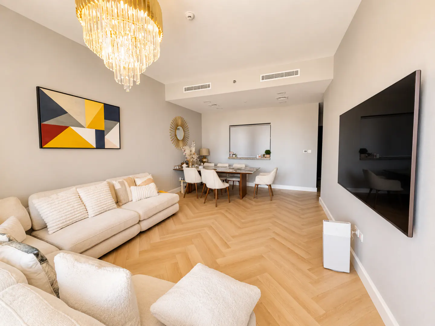 Modern living room with a cream sectional sofa, herringbone wood floors, a crystal chandelier, and a wall-mounted TV.