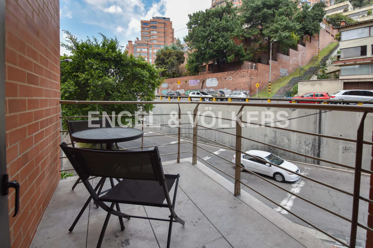 Balcony view with table and chairs overlooking a street with cars and buildings in the background.