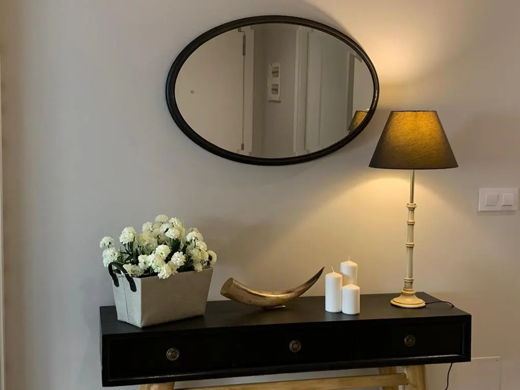 Entryway table with black drawers, topped with flowers, horn, and candles. Oval mirror hangs above, with a lamp to the right.