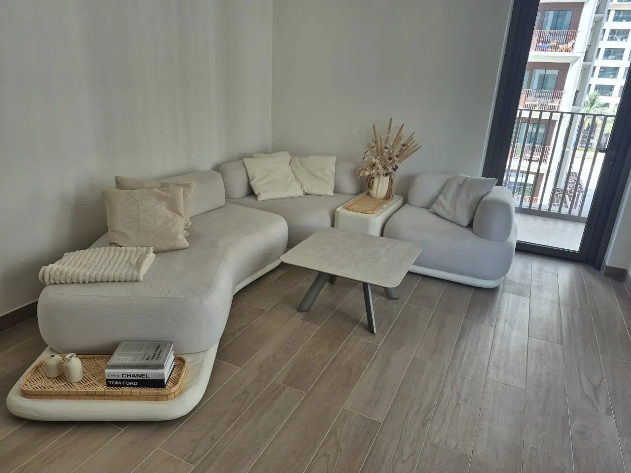 A modern living room features a curved white sofa with pillows, a square coffee table, and a balcony with a view of a high-rise building.